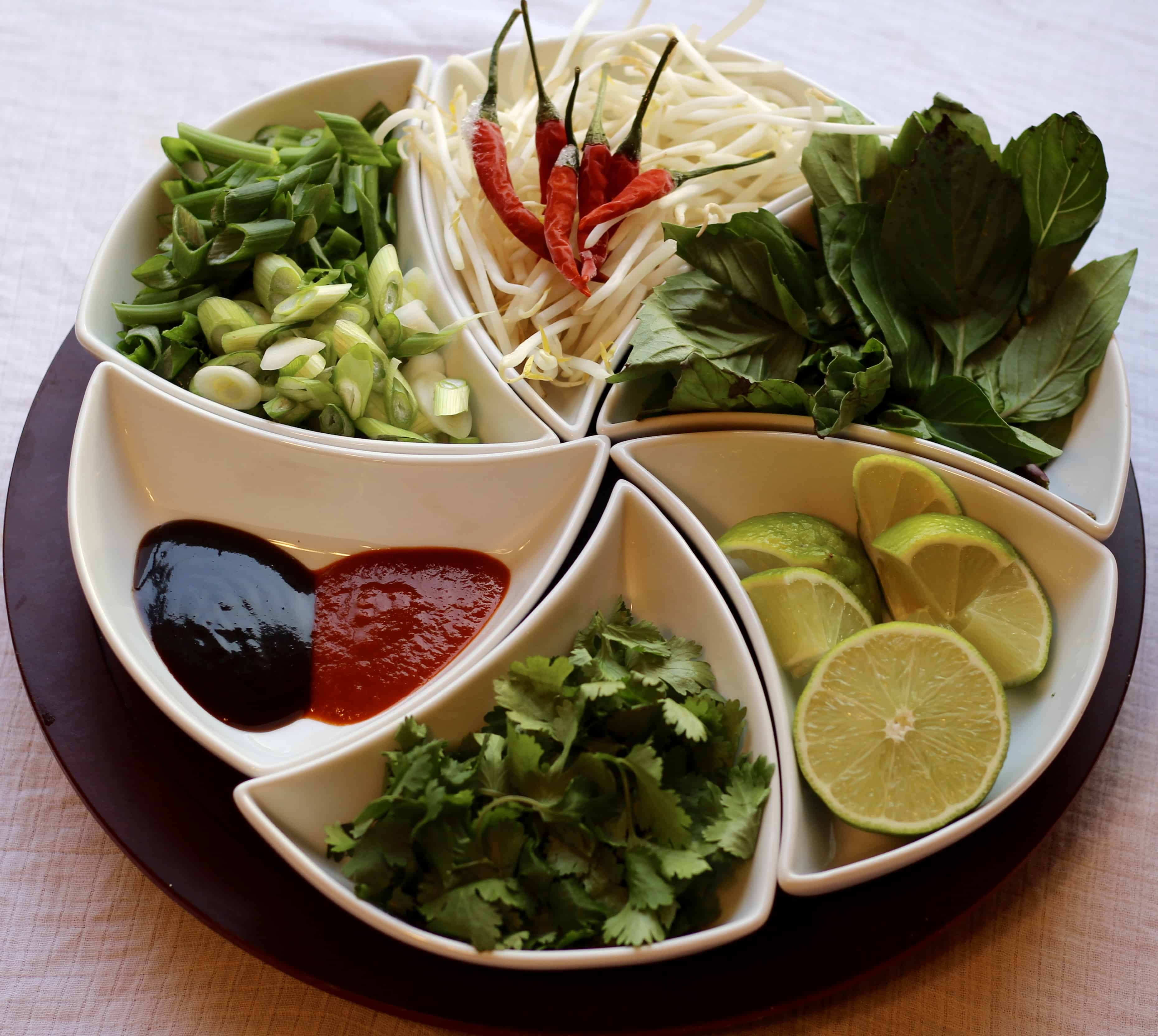 Pho toppings for homemade beef pho including thai basil, lime, green onion, bean sprout, hoisin sauce, and sriracha. 