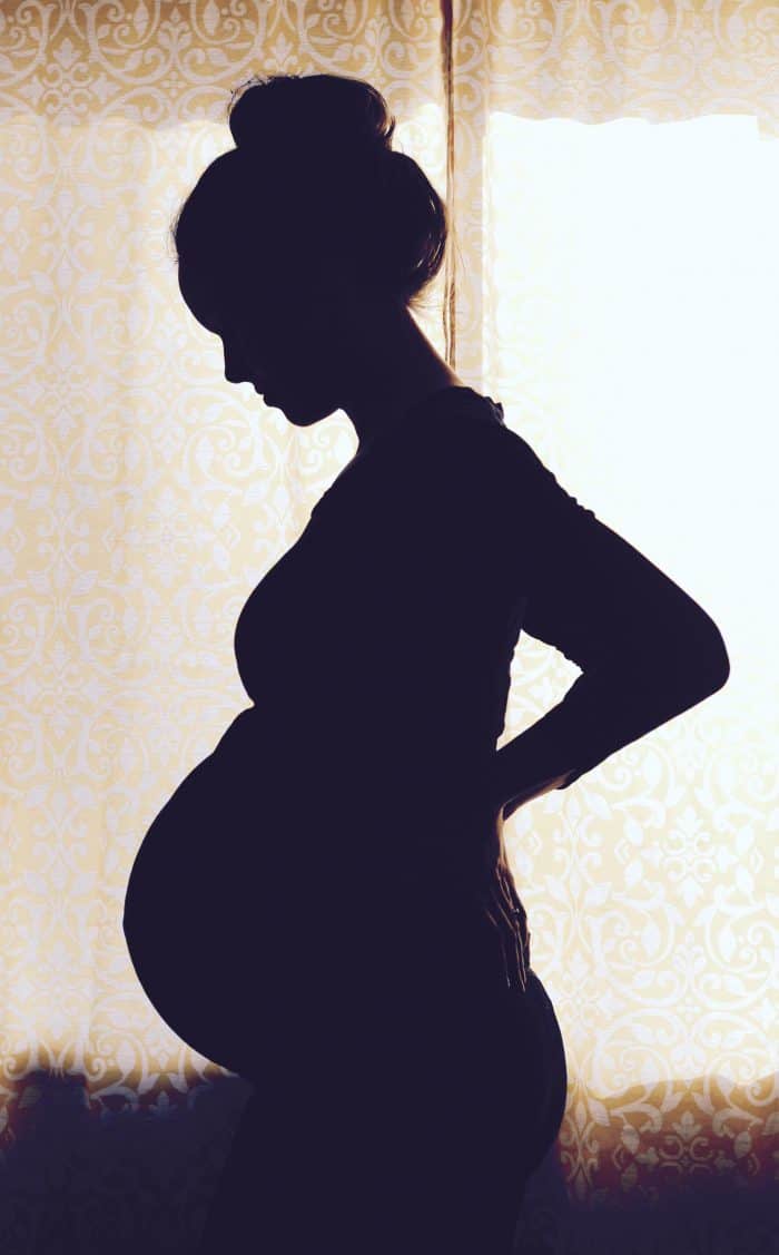How to take your own pregnancy silhouette of a woman standing in a window looking down as she uses an IPhone or DSLR camera at home