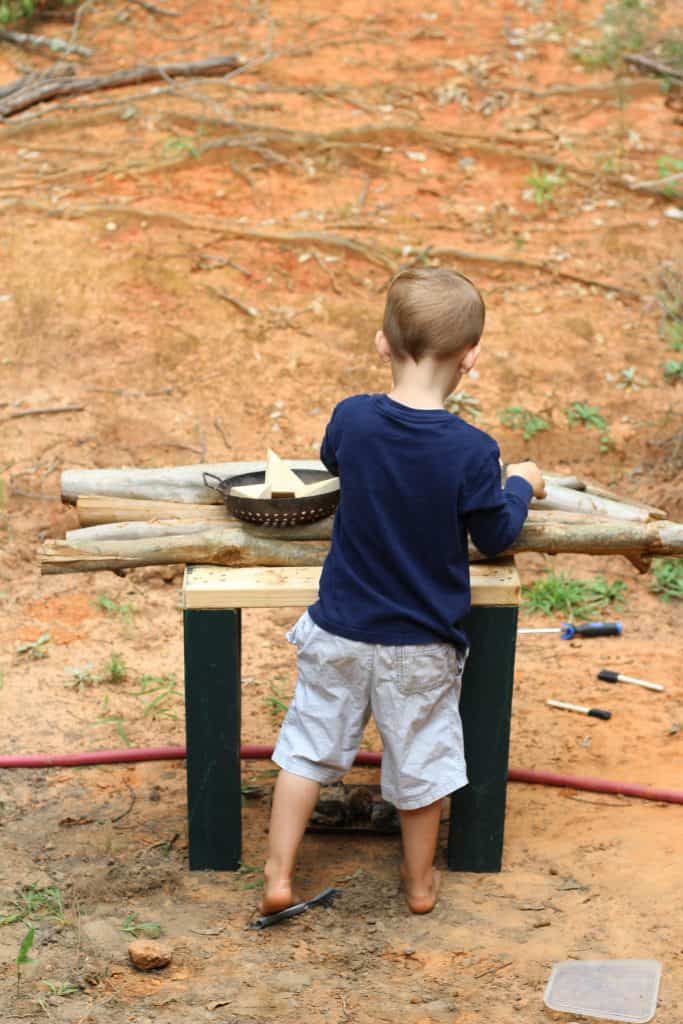 a little boy cooking in an outdoor play kitchen made of small wooden logs.