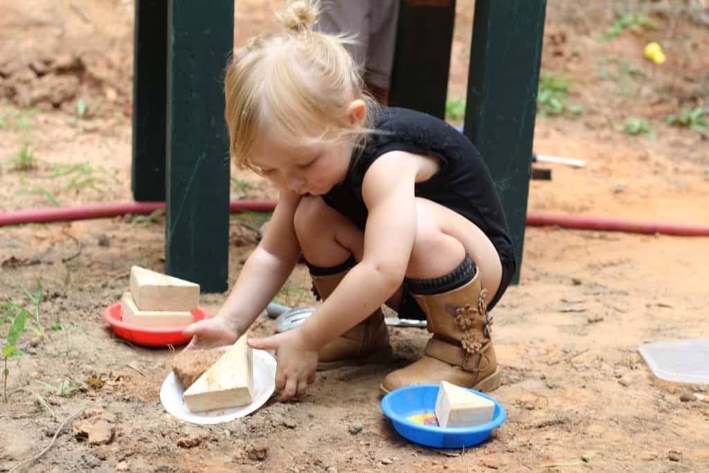 A toddler girl stacking wooden triangles on plastic plates in the dirt. 