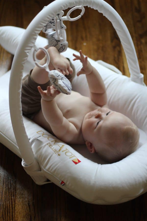 baby on his back playing with the toy arch above his white DockaTot 