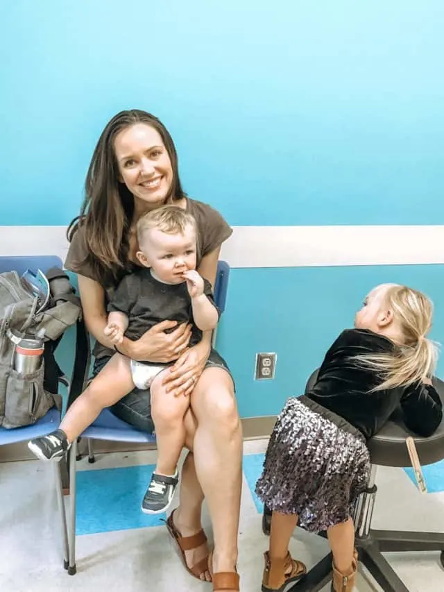 a mom holding her toddler while waiting to see the doctor, while a toddler spins on a chair in the waiting room