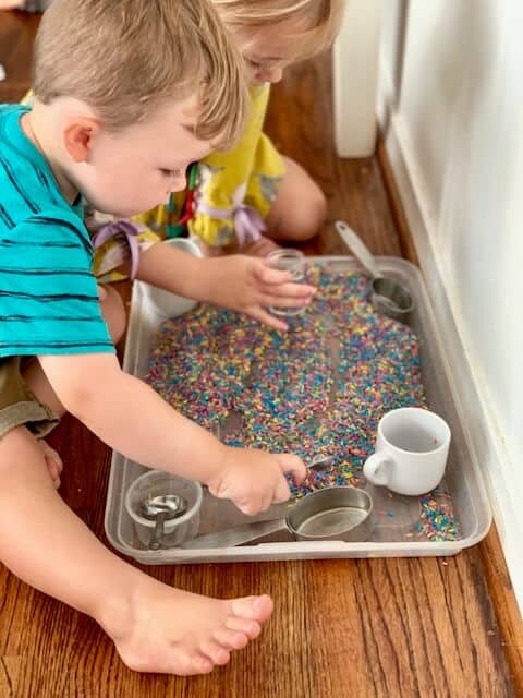 Two kids playing with colorful rice using scoops, small cups, and spoons. 