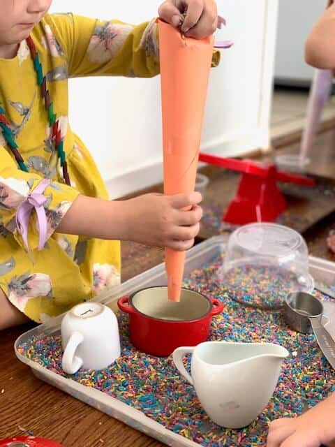 a little girl using a paper funnel with dyed rice in a pan