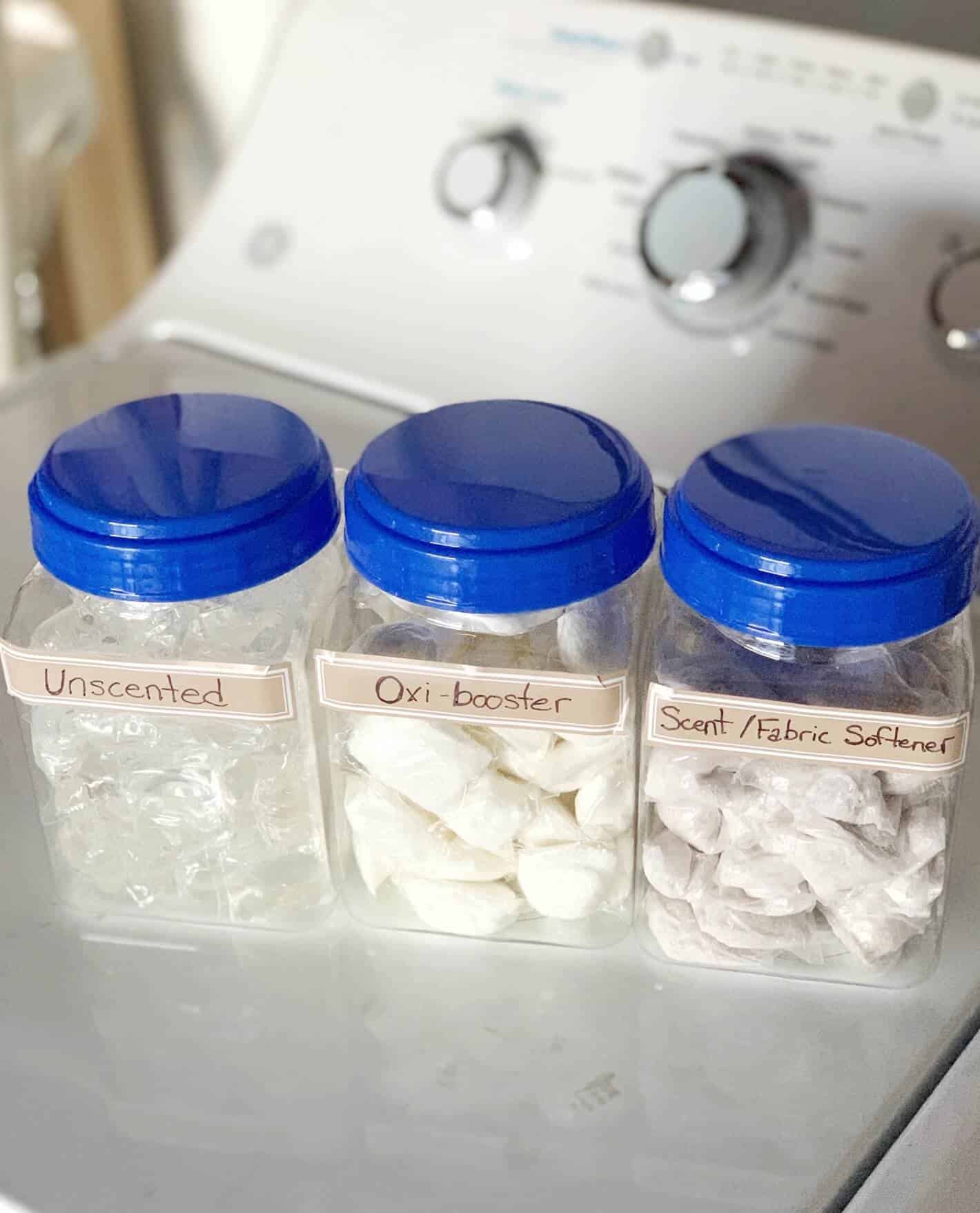 Dropps pods stored in 3 separate clear containers from the dollar store. One is labeled unscented, one is labeled oxi-booster, and one is labeled scented fabric softener. 