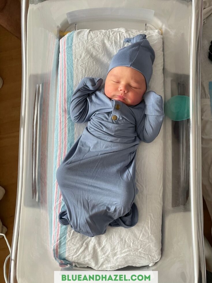 A newborn baby sleeping in the hospital bassinet wearing a blue hat and blue knotted onesie. 