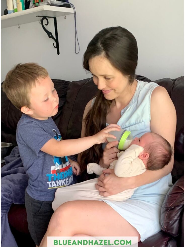 A mother feeding her newborn a bottle using the comotomo 5 oz wide neck bottle as her older boy helps by holding up the bottle