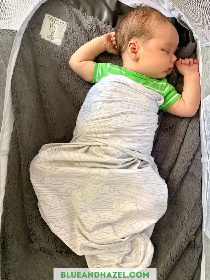 A newborn sleeping in a bassinet swaddled in a white cloud velcro swaddle