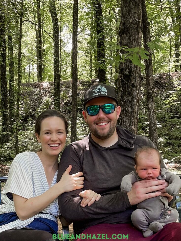 A husband and wife smiling in the woods while sitting and holding their newborn. 