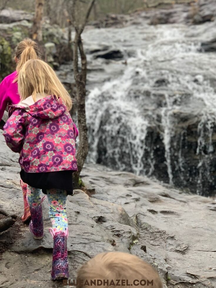 A little girl walking next to a small waterfall wearing rain boots