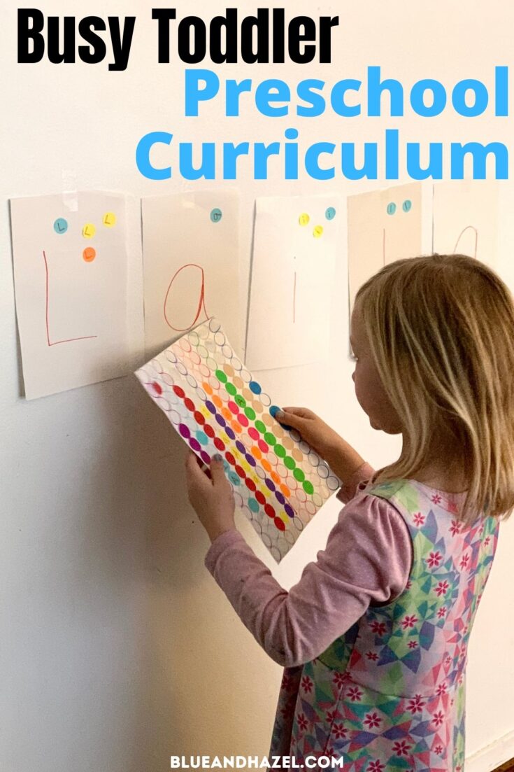 A toddler girl putting colorful dot stickers on paper on the wall as part of a preschool program by Busy Toddler. 