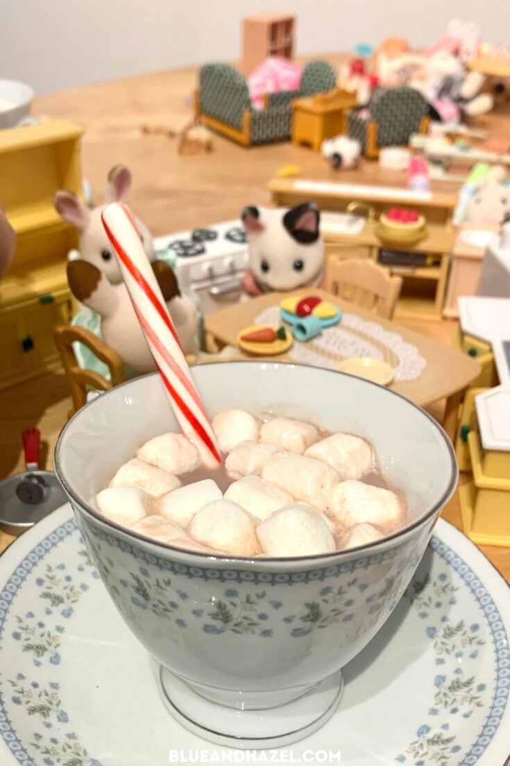 A tea cup full of cocoa with miini marshmallows and a candy cane, with Calico critters sprawled all over the table. 