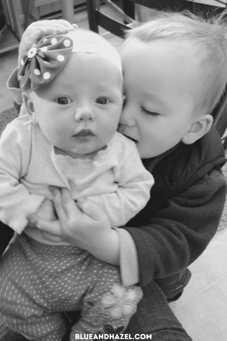 a black and white photo of a newborn baby girl being held by her toddler brother 18 months older. 