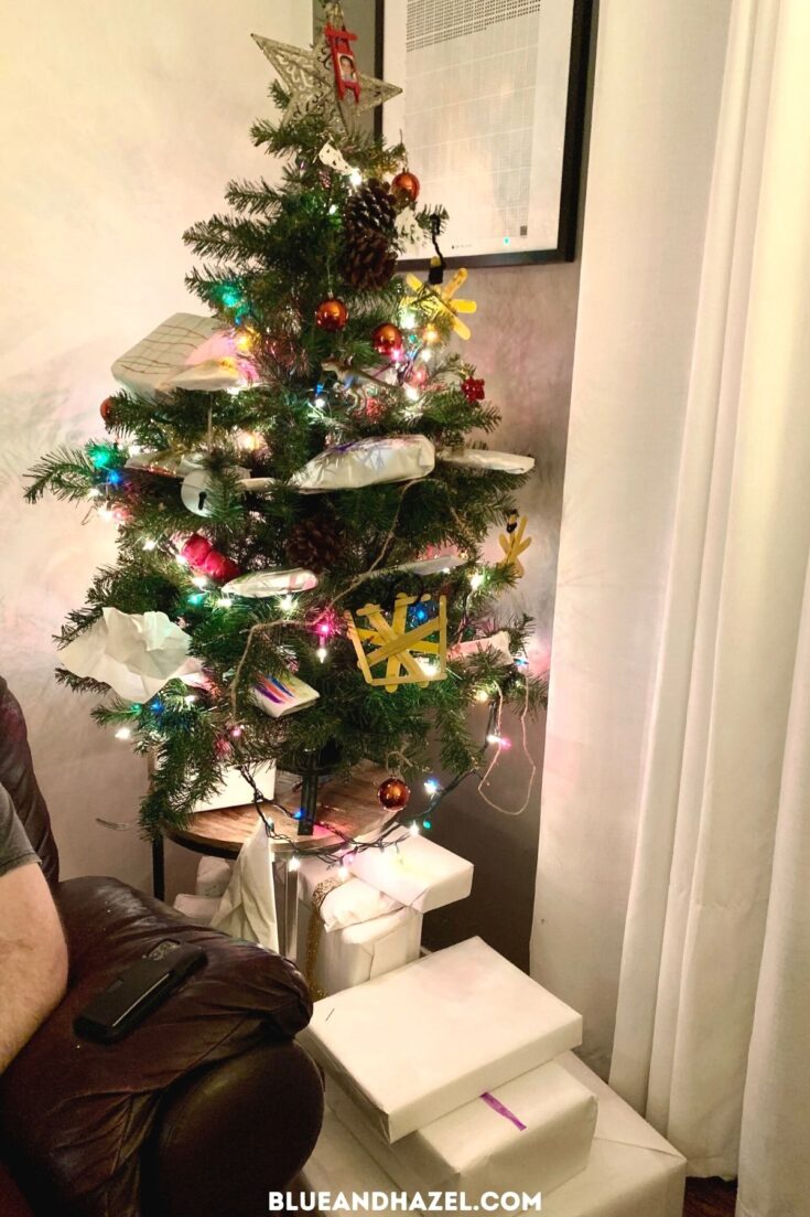 A small Christmas tree on a side table stuffed with homemade ornaments. Presents are tucked in the tree wrapped in white wrapping paper. 