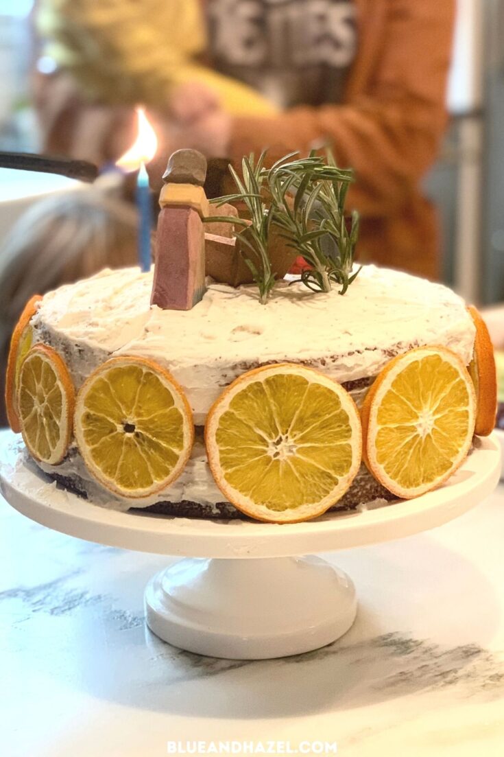 A white double layer round cake decorated with orange slices and topped with rosemary sprig and jesus in a manger. 