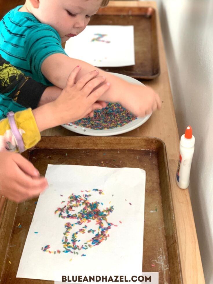 A toddler spreading glue on a white piece of paper and then sprinkling rainbow rice on top.