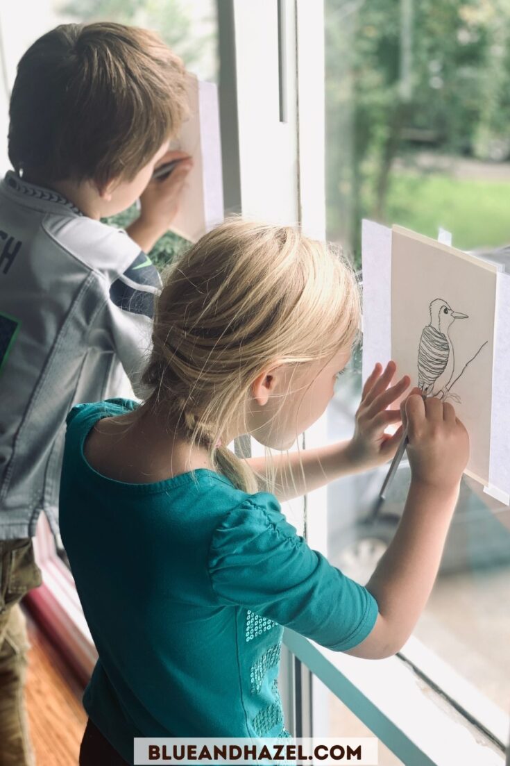 Two kids tracing a woodpecker on a window onto a piece of paper. 