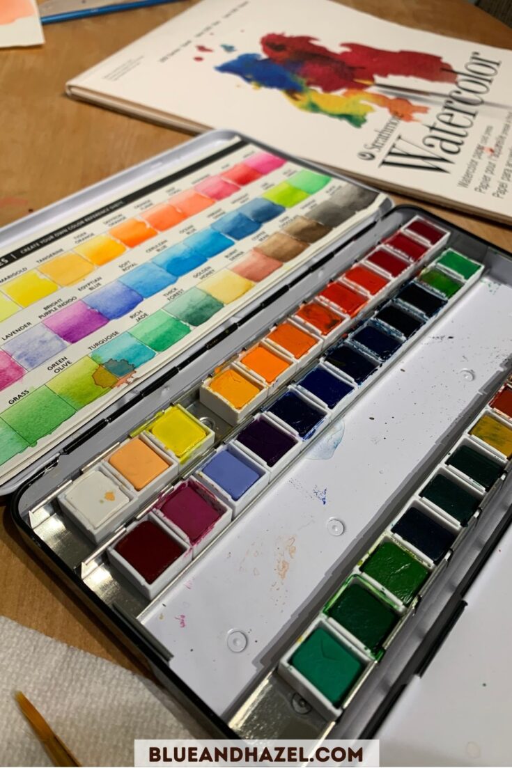 A watercolor paint set with 3 rows of colors, next to a pad of watercolor paper.