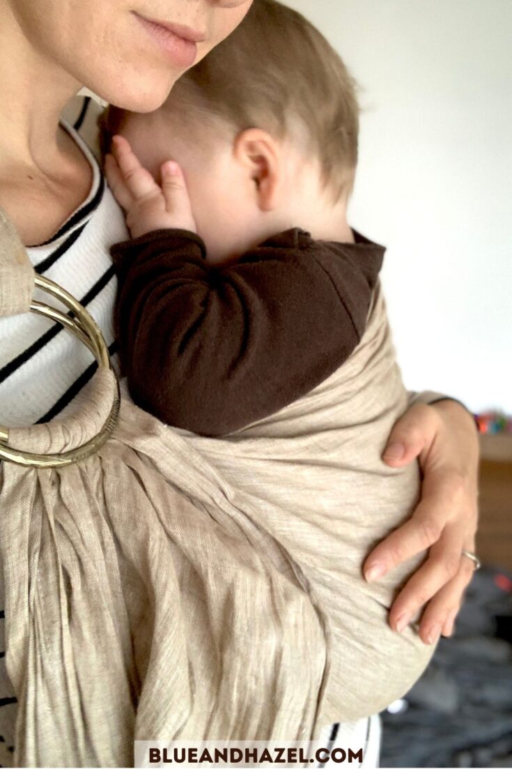 A close up of a mom holding her baby in a tan Lillebaby ring sling while the baby rubs his eyes. 