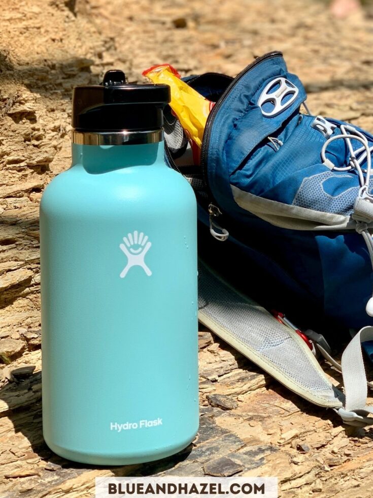 Hydro Flask 64 oz water bottle in Alpine next to a blue Osprey backpack on a rocky surface. 