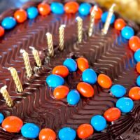 A round chocolate birthday cake with blue and orange m and m's around the edge with 9 gold candles