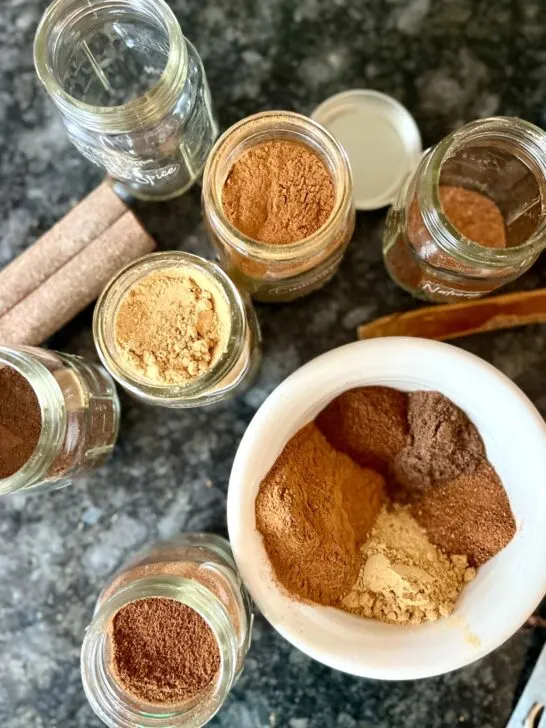 cinnamon, ginger, cloves, allspice, and nutmeg in open jars with all the spices being mixed in a small white bowl to create a pumpkin pie spice blend.