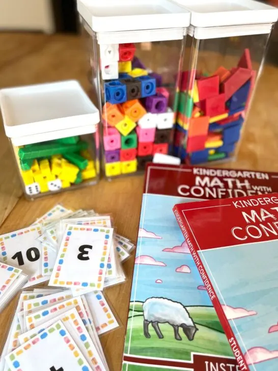Kindergarten math with confidence student book and instructor's manual on a kitchen table next to tubs of math manipulatives like math cubes, place value blocks, and colorful tangrams.