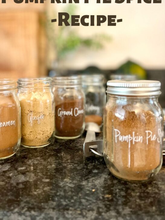 Homemade pumpkin pie spice in a 4 oz labeled jar with cloves, cinnamon, ginger, allspice, and nutmeg also in jars behind it.