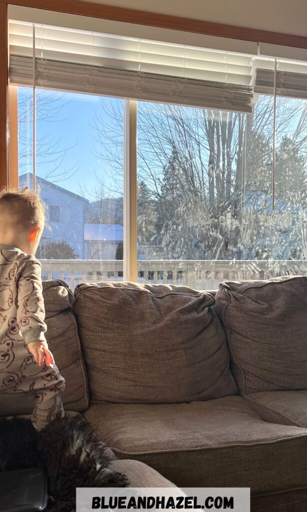 A dirty window with a toddler standing next to all the handprints on the window. 