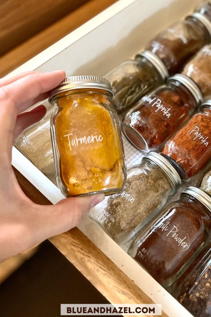 A hand holding a 4 oz mini glass jar of Turmeric spice with a drawer full of other spices in the background. 