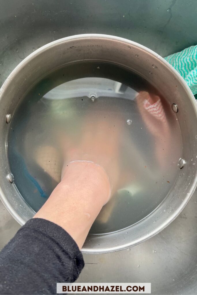 A hand reaching into murkey water filled with dirty, stinky Norwex microfiber cloths and Force Of Nature reusable paper towels.