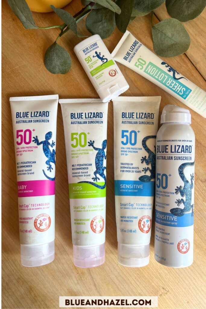 Blue Lizard sunscreen bottles on a wooden table including their SPF 50 spray, baby, sensitive, and kids SPF 50 lotions, sunscreen stick, and sheer sunscreen lotion. 